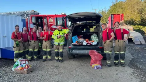 Clun Fire Station Eight firefighters in red t-shirts and unifom trousers smile as they stand in front of an open car boot containing drinks and snacks, with fire engines in the background.