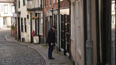 Shaun Whitmore/BBC The cobbled street of Elm Hill. The street is lined with independent shops and businesses. Outside one business a man is wearing a flat cap and is staring into one of its windows.