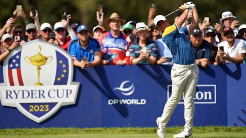 PA Media Action shot of Rory McIlroy playing a tee shot at the 2023 Ryder Cup. He is wearing the Team Europe outfit of a blue and yellow shirt and cream trousers. A throng of excited-looking fans stand behind him, some of whom have camera phones in hand.