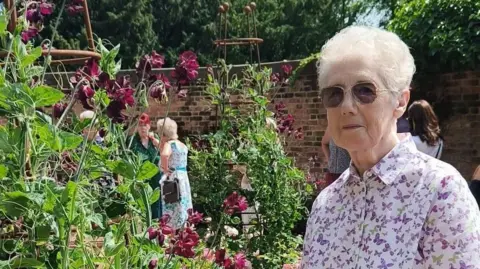 A woman, Audrey Morris, stands in a walled garden near flowers in the sunshine.
