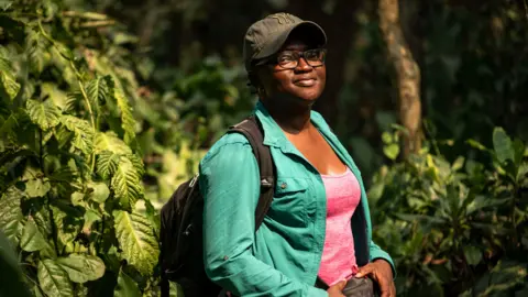 Etinosa Yvonne/Goldman Environmental Prize Iroro Tanshi smiling and wearing a dark cap and glasses. She is wearing a pink tshirt and a green open shirt on top. She has a rucksack on her back, and is standing surrounded by trees.