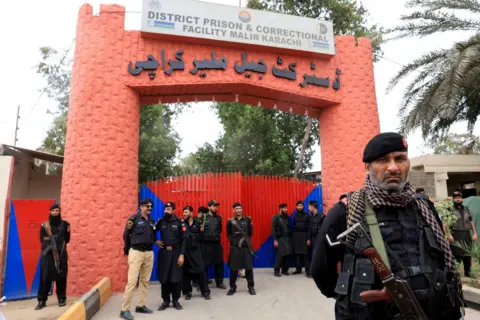 Reuters Police officers in uniform with rifles strapped around them stand outside large red and blue gates with a sign above them which reads "District Prison & Correctional Facility Malir Karachi".