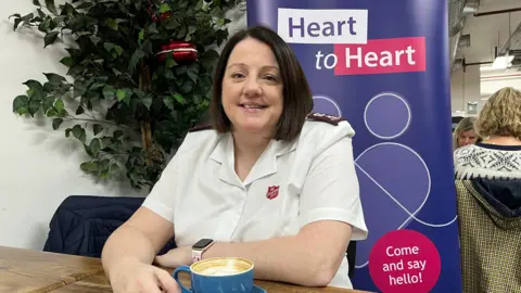 Major Ali Friday from The Salvation Army sits with a coffee at The Canterbury Bakery with a pull-up banner behind her which reads 'Heart to Heart' and 'Come and say hello!'
