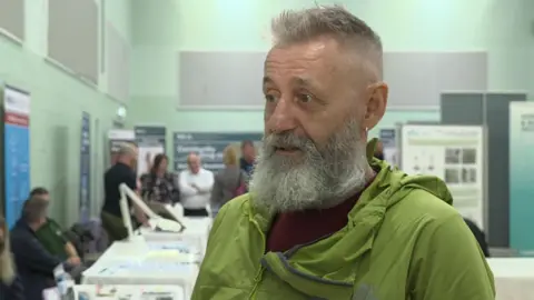 A man with a grey beard and green jacket. He is wearing a red t-shirt. There are stalls and posters behind him.