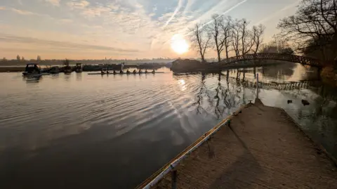 Lucie Johnson The sun glints off the water of the river. In the distance are narrowboats, a metal footbridge, and a boat with eight female rowers and what looks like a cox on the end.