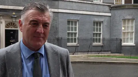 Ian Byrne MP has grey hair and is wearing a blue shirt, grey suit and tie. He is standing in the rain across the road from 10 Downing Street.