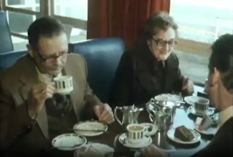 Screen grab from a 1970s TV report showing three people around a table in a motorway service station of the time. China cups and silver teapots are visible on the table, with blue leatherette seats.