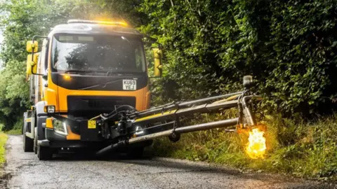 The 'dragon patcher' out and about in Devon. It is a large yellow truck with an extendable metal arm. At the end of the arm you can see a flame firing from a cannon-like machine part. The truck has a yellow flashing light on its roof. There is grassland either side of the road.