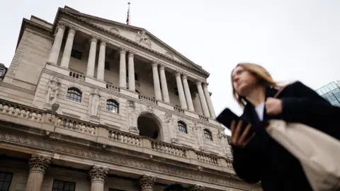 The Bank of England is pictured against a grey sky with people walking in front holding their phones, in Bank, London in December.
