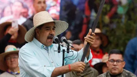 Reuters Maduro, speaking into a microphone and holding an old sword. He's wearing a light blue shirt and a straw hat.