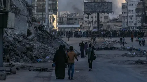 EPA Um homem e uma mulher palestina andam por uma rua cheia de detritos e escombros na cidade de Gaza. À distância, há uma multidão de pessoas, prédios destruídos e fumaça onda no céu.