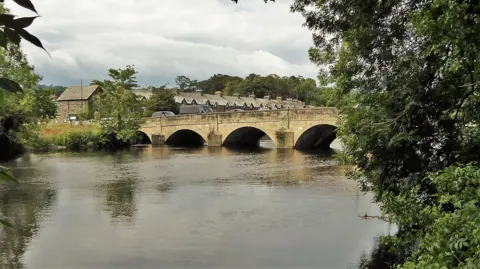 A stone bridge crossing a wide river. There is traffic queuing on the bridge. 