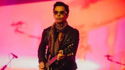 Kelly Jones of Stereophonics performing on the main stage at the Isle of Wight Festival. He has short dark hair and wears sunglasses, a black leather jacket and a dark red scarf. He's playing an electric guitar.