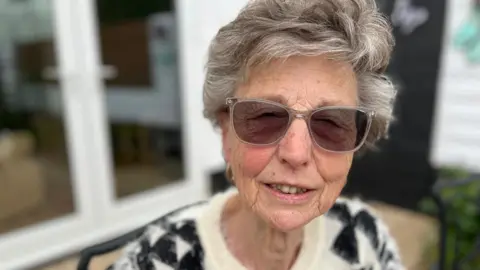 Val has short grey hair and is wearing tinted glasses. She is sat in a garden, wearing a black and white jumper
