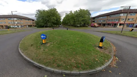 Conniburrow Boulevard in Milton Keynes, taken from a Google Streetview camera. In the foreground is a grassed mini roundabout. The road continues straight into the background, with flats and trees on both sides. Underneath one flat is a kebab shop.