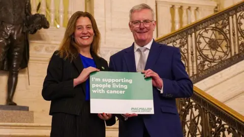 Chief Executive of Macmillan Cancer Support Gemma Peters, and Health Minister Mike Nesbitt stand on the steps in Stormont's Great Hall and smile at the camera. They are holding a small board from Macmillan cancer support which states: 'I'm supporting people living with cancer'