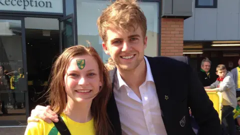 Roberth Kristiansen Emmy wearing a Norwich City shirt and with the club badge painted on her forehead standing next to footballer Patrick Bamford who us wearing a black suit and has his arm around her. They're standing outside Carrow Road