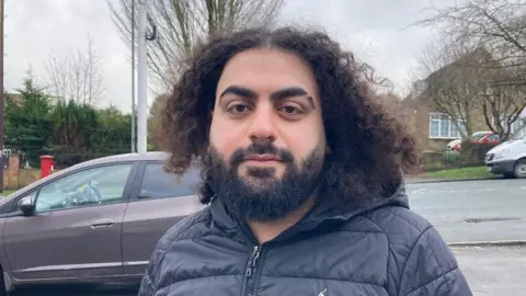 A man with curly hair wearing a black coat standing outside with the yellow camera on a metal pole in the background