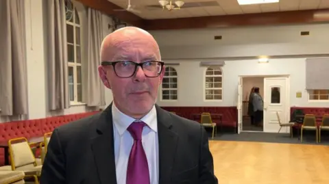 MP Matt Western wearing dark glasses, a black suit and a purple tie in a village hall