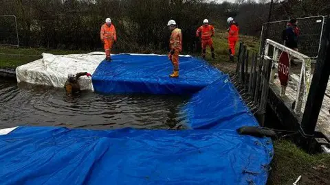 Men in high visibility orange outfits on the edge of a canal building a dam covered with a blue tarpaulin. Next to them is a small bridge.