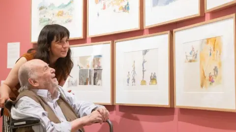 Quentin Blake Sir Quentin looks at a series of drawings in frames on a wall with a joyful expression. A woman with brown hair stands behind him also looking at the frames.