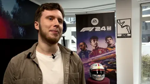 George is wearing a brown shirt which is open with a white T-shirt underneath. He has brown hair and is stood next to some F1 promotional items and a race helmet.