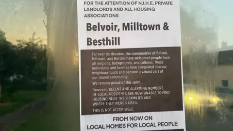 A poster on a glass wall of a bus stop. It is brown and white in colour and has text on it which says, for the attention of N.I.H.E, private landlords and all housing associations - Belvoir. Milltown & Besthill - from now on local homes for local people. 