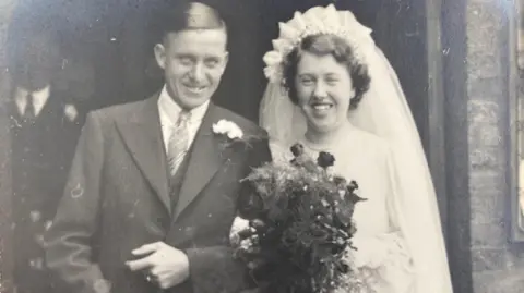 Supplied Leslie and Doreen on their wedding day, in a black and white image. He is wearing a suit, with a flower in his lapel, Doreen has a veil and head dress on, a white wedding dress and is holding a bouquet of flowers.