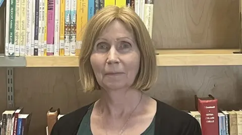 Aodhán Gregory Linda Ervine, a woman with blonde hair styled in a bob, looks directly at the camera. She is standingi front of a wooden bookshelf. She is wearing a black cardigan over a dark green top. 