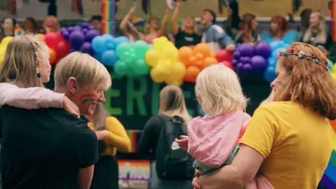 Curious North Productions A crowd gathers at a colourful outdoor Pride event, with people standing beneath a large arch of rainbow balloons. In the foreground, two female adults hold young blonde-haired children while looking toward the lively scene ahead. Many attendees wear bright clothing, and pride flags and decorations and balloons are visible throughout the background.