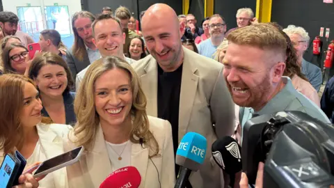 A group of people are standing together smiling. Mics are being held up towards them. The room is full of people. 