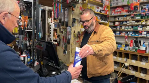 John Cox wears a sand-coloured jacket, dark sweater and trousers. He hands a customer a tube of instant filler. The customer wears glasses and a dark blue jacket. Hardware goods are visible in the background. 