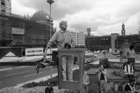 SSPL/Getty Images A black and white photo of a man standing in the bucket of a crane above a street in a town centre with a wide road and large buildings behind him. He has a mop of grey hair and wears glasses, a jumper and jeans with a black camera hanging from around his neck. A group of people including a man taking a photograph, a standing beneath the bucket. A police officer stands to the left.