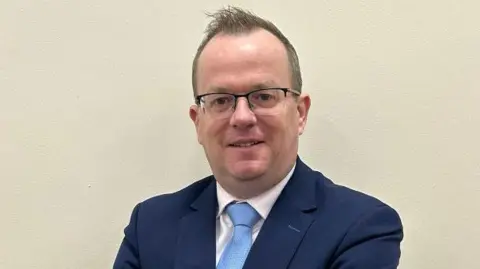 Regent House School A man with dark hair, wearing black glassses, pictured against a beige wall. He is wearing a navy suit jacket, blue tie and white shirt.