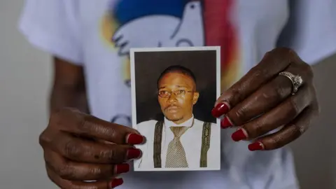 AP is close to the hands of the woman, with the reading of the nail reading, holding a small image of the Bwana Chui Bin Kositi. In it, stare at the foreground and appears dangerous. He wears a glass -wire glass, a silver and black tie, black arches and a white shirt. A woman's shirt, which shows the design of the peace dove, can be seen in the background.