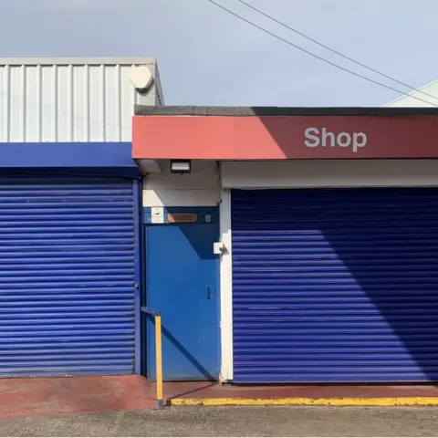 Tom Hicks Two blue industrial units have horizontal corrugated iron shutters. In between two units there is a blue door. Above the left hand unit is a panel with vertical white corrugated iron. Above the right shutter is a red panel with Shop written in white lettering. 