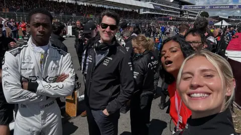 A selfie photo by Callie Cooke with her smiling alongside Simone Ashley wearing red, Joseph Kosinski in black and Damson Idris in a white motor-racing suit. They are in the pit lane at Silverstone while people work behind them.