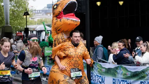 A runner in a dinosaur costume runs in the London Marathaon with crowds clapping in the background.