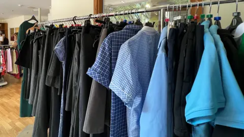 The image shows clothing racks inside a charity shop displaying school uniforms and other clothes, with shirts, skirts and trousers hanging on rails in a bright indoor space.
