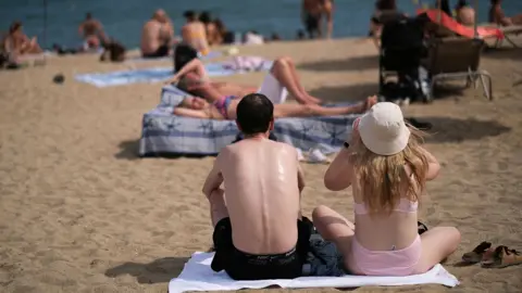 Reuters Two people sit on a beach, one in a bucket hat, looking at the sea with their backs turned to the camera and crowds of people in the background, on a sunny day in Barcelona in August 2024.