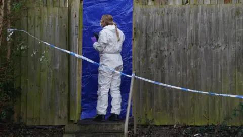 PA A forensic officer in a white suit standing next to a blue tent. She is behind police tape in a garden obscured by a fence.
