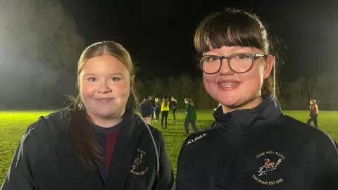ELLEN KNIGHT/BBC Eden and Jess stood side by side, both smiling and looking into the camera. They're stood on a flood-lit rugby pitch, which has green grass, a dark night sky, and is bordered by trees. Both girls are wearing black zip-up sports jackets, which have a logo on the chest that features a ram, and 'Clee Hill RUFC, The Rams Est 1998' written on it. 