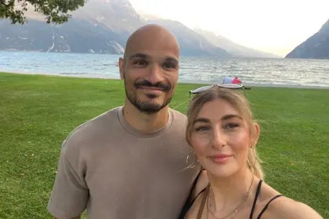 Emma Giannuzzi A selfie of a man and woman smiling while standing next to a lake or the sea, with some mountains behind them. The man is balding and has a black goatee, and wears a brown t-shirt. The woman is blonde and has a black strappy top on. It is a head and shoulders shot of them.