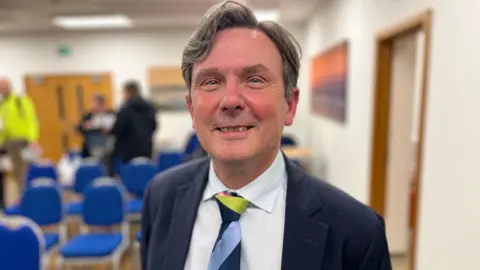 BBC Deputy Andy Sloan - A man with grey and black wavy hair looking towards the camera smiling. He's wearing a blue suit, with a white shirt and a striped tie with green, blue and navy stripes. 