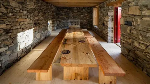 National Trust for Scotland A long wooden table inside the cottage. There is stone brick work along all four walls, with the red door open allowing in the daylight. Two long wooden benches are on either side of the table. 