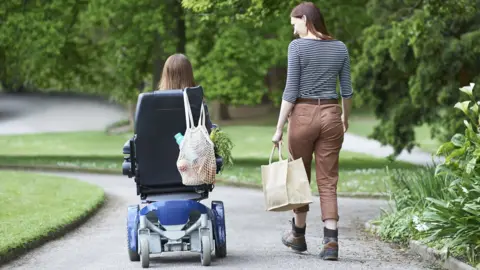Seen from behind, a woman holding a shopping bag walks along a garden path alongside a long-haired person in a motorised wheelchair, their own shopping bag hung over the top corner of the chair