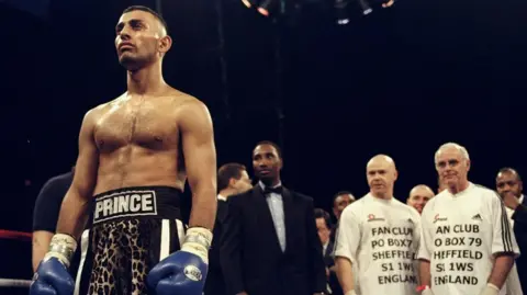Getty Images Prince Naseem Hamed (left) stands focused in the ring. He wears his leopard print shorts with prince logo on the waistband. Brendan Ingle and another assistant stand at the back next to the referee, wearing white shirts with the fan club postal address on their front.