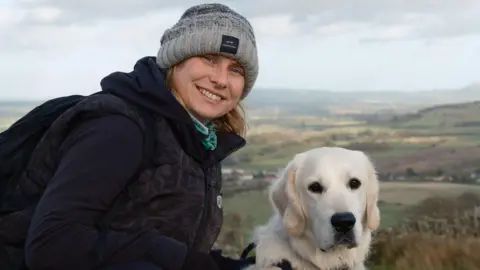 SUPPLIED Natalie in grey beanie in dark hoodie and body warmer with golden retriever dog stood in front of hillside landscape