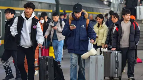Getty Images Young travelers pull their suitcases and check their phones while walking along the platform of Fuyang Railway Station. 