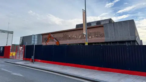 A large black barrier and red gate is erected in front of The Potteries Museum and Art Gallery. The arm of a digger can be seen behind the barrier.